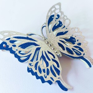 Peut inclure: Une d&eacute;coupe de papier en forme de papillon avec des paillettes argent&eacute;es et bleues et des d&eacute;coupes complexes. Le papillon est compos&eacute; de deux couches, une couche de paillettes argent&eacute;es sur une couche de paillettes bleues.