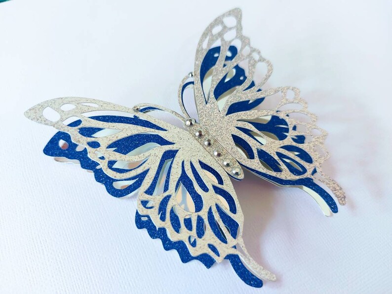 Peut inclure: Une d&eacute;coupe de papier en forme de papillon avec des paillettes argent&eacute;es et bleues et des d&eacute;coupes complexes. Le papillon est compos&eacute; de deux couches, une couche de paillettes argent&eacute;es sur une couche de paillettes bleues.