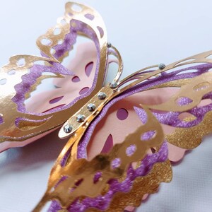May include: A close-up of a layered butterfly decoration. The butterfly features gold, glittery purple, and light pink paper cutouts. Rhinestones are placed along the center of the butterfly. The wings have intricate cut-out designs.