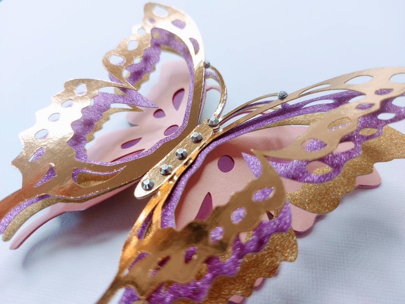 May include: A close-up of a layered butterfly decoration. The butterfly features gold, glittery purple, and light pink paper cutouts. Rhinestones are placed along the center of the butterfly. The wings have intricate cut-out designs.