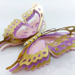 May include: A layered paper butterfly in gold, lavender, and pink. The butterfly has intricate cut-out details on its wings and is embellished with small, silver-colored rhinestones. The wings have a glittery texture, and the overall design is three-dimensional.