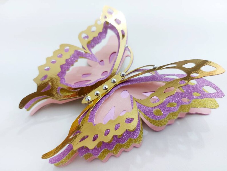 May include: A layered paper butterfly in gold, lavender, and pink. The butterfly has intricate cut-out details on its wings and is embellished with small, silver-colored rhinestones. The wings have a glittery texture, and the overall design is three-dimensional.