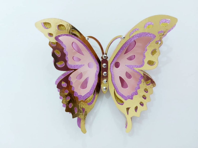 May include: A decorative butterfly with layered wings. The top layer is gold with brown spots, the middle layer is pink, and the edges are outlined with purple glitter. The butterfly has a row of rhinestones down the center.