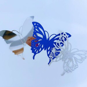 Peut inclure: Trois papillons d&eacute;coratifs en papier. Un papillon est argent&eacute;, un autre est bleu et le dernier est blanc avec des paillettes.