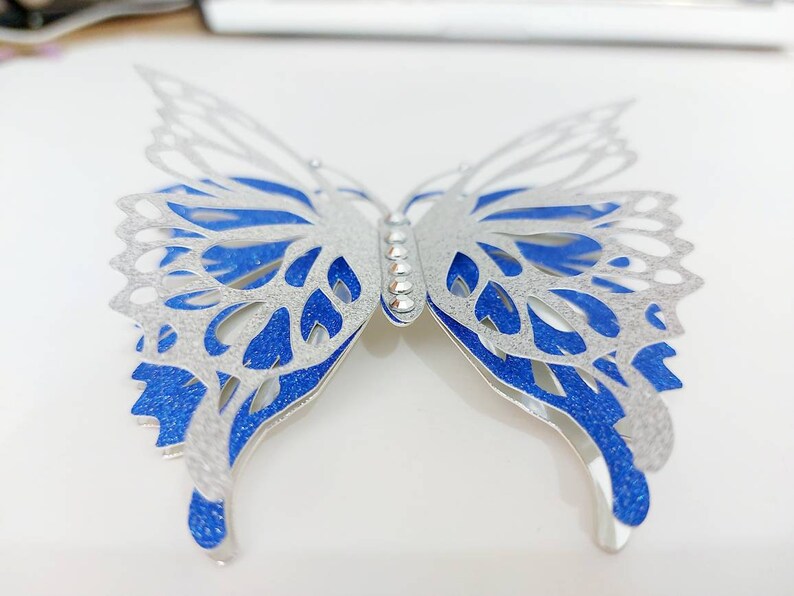 Peut inclure: Une d&eacute;coration murale en forme de papillon argent&eacute;e et bleue avec une finition scintillante. Le papillon a des d&eacute;coupes complexes et une rang&eacute;e de strass au centre.