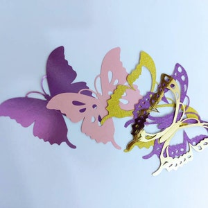 May include: A collection of paper butterflies in various colors. The butterflies are in shades of purple, pink, and gold. The butterflies are cut out with intricate details, and are arranged on a white background.