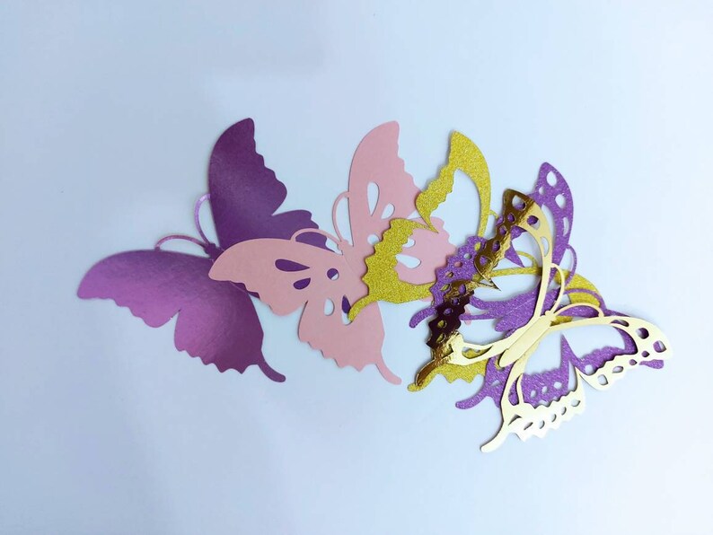 May include: A collection of paper butterflies in various colors. The butterflies are in shades of purple, pink, and gold. The butterflies are cut out with intricate details, and are arranged on a white background.