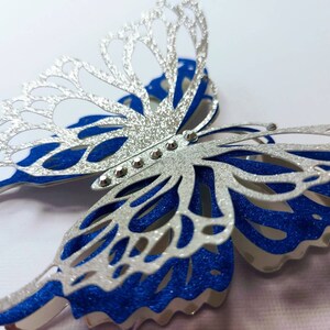 Peut inclure: Un papillon en paillettes argent&eacute;es et bleues avec un design 3D. Le papillon a un corps en paillettes argent&eacute;es et des ailes en paillettes bleues. Le papillon est d&eacute;cor&eacute; de strass argent&eacute;s.