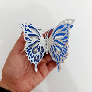 Peut inclure: Une d&eacute;coration en forme de papillon en paillettes argent&eacute;es et bleues avec des d&eacute;coupes complexes. Le papillon est tenu dans une main.