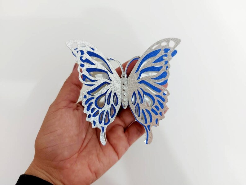 Peut inclure: Une d&eacute;coration en forme de papillon en paillettes argent&eacute;es et bleues avec des d&eacute;coupes complexes. Le papillon est tenu dans une main.