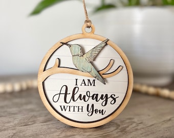 May include: A round wooden ornament with a hummingbird design. The hummingbird is perched on a branch, with the words "I AM Always WITH You" in black script. The ornament has a natural wood frame and a twine hanger.