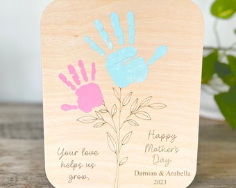 May include: Wooden plaque with a light wood grain finish. The plaque features a hand-drawn floral design with two handprints in pink and blue. Text reads "Happy Mother's Day" and "Your love helps us grow."