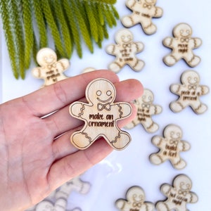 May include: Wooden gingerbread man ornament with the text "make an ornament" in a handwritten font. Other gingerbread men with various holiday phrases are scattered around. The ornaments are light brown and have a smiling face.