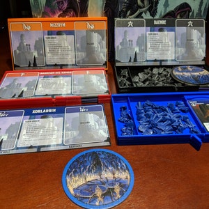 May include: A tabletop game setup featuring various components. There are four card holders in orange, black, and red, each labeled with names like "Mizzyrm" and "Baenre." A blue tray holds game pieces and cards. A circular game piece is also visible.