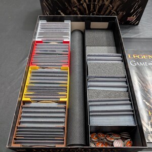 May include: A black board game box with internal organizers. The box contains stacks of cards in red, yellow, and brown dividers, along with a black cylindrical object and game tokens. The box is labelled "Game of Thrones."