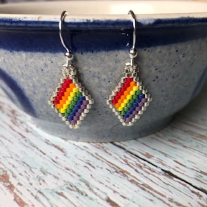 May include: A pair of rainbow-coloured beaded earrings with silver hooks. The earrings are shaped like diamonds and feature a rainbow pattern of red, orange, yellow, green, blue, indigo, and violet beads.