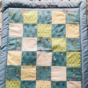May include: A patchwork baby quilt with a blue and white polka dot border. The quilt features squares of light blue fabric with bunnies, green fabric with leaves, and white fabric. The words "Sweet Dreams" are embroidered on the top.