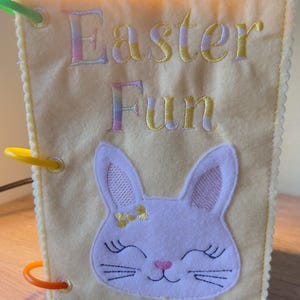 May include: A yellow felt book with the words "Easter Fun" embroidered in pastel shades. A white bunny face with closed eyes and a pink nose is appliquéd on the front. The book has colourful plastic rings and a scalloped edge.