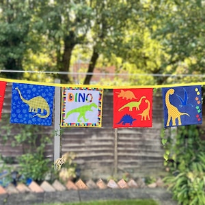 May include: A dinosaur-themed bunting with six square flags. Each flag displays a different dinosaur design in various colours, including red, blue, and yellow. The flags are strung on a yellow cord, creating a decorative banner.