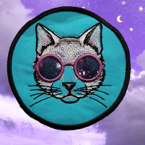 May include: Embroidered patch featuring a cat's face wearing pink sunglasses with a galaxy design. The cat is gray and white, set against a teal background with a black border. The patch is round and set against a purple and blue sky.