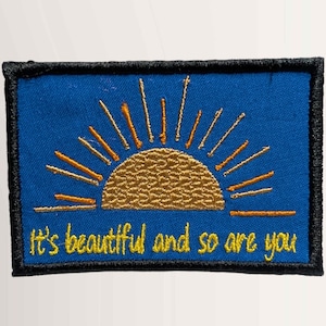 May include: Embroidered patch with a blue background, featuring a golden sun design with rays. The text "It's beautiful and so are you" is in yellow. The patch has a black border.