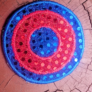 May include: Circular patch with concentric rings of blue and red sequins. The outer ring is blue with blue sequins, followed by a red ring with red sequins, and a blue center. The patch is embroidered with a blue border.