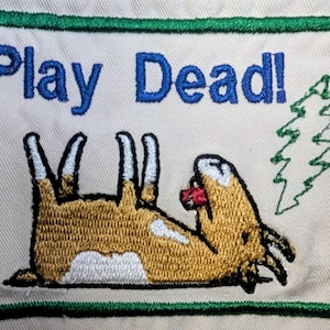 May include: Embroidered patch with the words "Play Dead!" in blue above a cartoon animal playing dead. The animal is brown and white with a red object in its mouth. A green tree is embroidered on the right.
