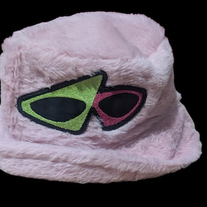 May include: A light pink, fuzzy bucket hat with an embroidered design. The design features two overlapping, stylized glasses shapes, one green and one pink, with black centers. The hat is set against a black background.