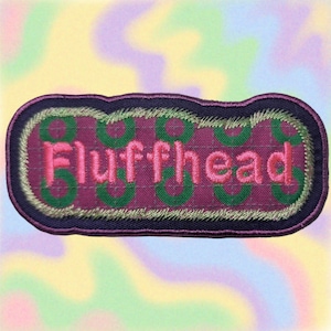 May include: Embroidered patch with the word "Fluffhead" in pink, set against a purple background with green circular accents. The patch has a dark purple border and gold stitching, perfect for jackets or bags.
