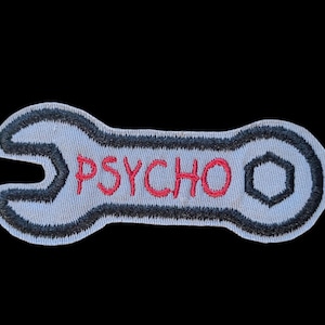 May include: Embroidered patch in the shape of a wrench, light blue with black trim. The word "PSYCHO" is embroidered in red across the center. The patch is set against a black background.