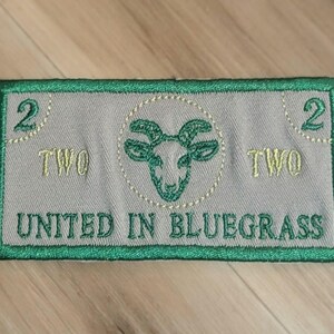 May include: Embroidered patch in the shape of a two-dollar bill. The patch is gray with green and yellow embroidery. It features a goat head, the word "TWO", and the text "UNITED IN BLUEGRASS".