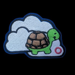 May include: Embroidered patch featuring a cartoon turtle with a green head and legs, and a brown shell. The turtle is set against a white cloud with a blue outline. A small pink circle is visible.
