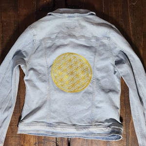 Puede incluir: Chaqueta vaquera azul claro con un diseño dorado bordado de la Flor de la Vida en la espalda. La chaqueta tiene un cuello clásico, cierre de botones en la parte delantera y mangas largas. El bordado dorado añade un elemento decorativo.