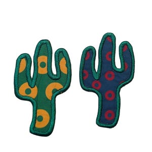 May include: Two cactus-shaped patches. One is green with orange circles, the other is navy blue with red circles. Both have green embroidered outlines. These patches are ideal for adding a unique touch to clothing or accessories.