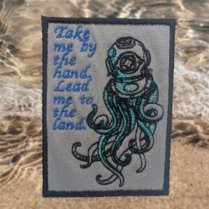 May include: A grey rectangular patch with a dark blue border. It features a teal and black illustration of a vintage diving helmet with tentacles and the text "Take me by the hand, Lead me to the land" in blue.