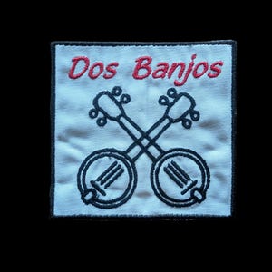 May include: Embroidered patch featuring two crossed banjos in black on a white background. The text "Dos Banjos" is stitched in red above the instruments. The patch has a black border.