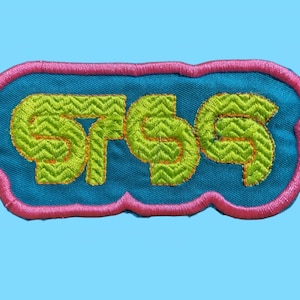 May include: Embroidered patch with the word "SASS" in lime green, outlined in gold, on a blue background. The patch has a pink border and a unique shape. The background is a light blue.