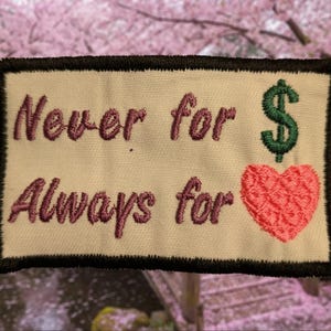 May include: Embroidered patch with the text "Never for" above "Always for". A green dollar sign is on the right, above a pink heart. The patch has a black border and is on a cream-colored background.