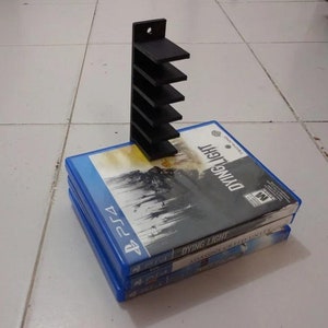 May include: A black plastic video game storage stand with five slots for holding video game cases. The stand is holding a stack of three video game cases, all with blue covers. The top case has the title "Dying Light" printed on it.