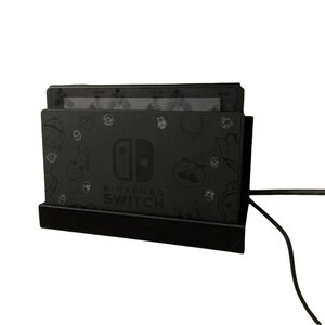 May include: A black Nintendo Switch console charging dock with a black power cord. The dock has a black and white design with a Nintendo Switch logo and the words "Nintendo Switch" printed on it.