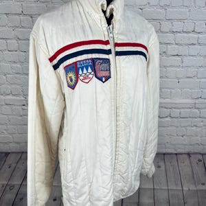 May include: Cream-colored quilted jacket with a high collar and a full-length zipper. The jacket features a horizontal stripe in red, white, and blue across the chest, along with three embroidered patches. The patches include text and various designs.