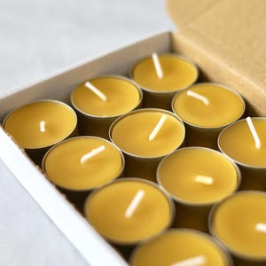 Può includere: Una scatola di lumini. Le candele sono di colore giallo caldo, con uno stoppino bianco al centro. Le candele sono in piccoli contenitori rotondi di metallo. La scatola è di cartone.
