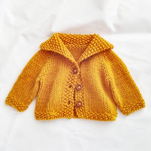 May include: Mustard yellow knitted baby cardigan with a shawl collar and three brown buttons. The handmade sweater has long sleeves and a textured knit pattern. A cozy and warm garment for a baby.
