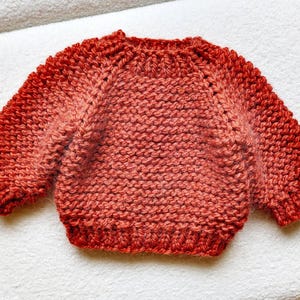May include: A hand-knitted, coral-colored sweater with a crew neck and long sleeves. The sweater features a textured knit stitch and a gradient effect, transitioning from a lighter coral to a deeper red at the cuffs and hem. The garment is displayed on a white surface.
