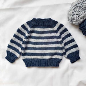 Könnte beinhalten: Ein gestrickter Pullover mit blauen und grauen horizontalen Streifen. Der Pullover hat einen Rundhalsausschnitt und lange Ärmel. Das Garn ist im Hintergrund sichtbar. Der Pullover ist aus weichem Garn gefertigt.