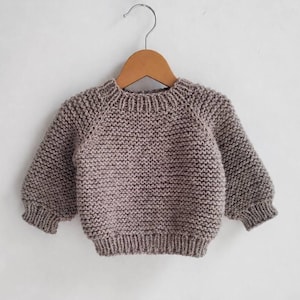 May include: A taupe-colored baby sweater, hand-knitted with a textured stitch. The sweater features long sleeves, a ribbed hem, and a crew neck. It is displayed on a wooden hanger against a plain white background. The sweater is designed for infants.