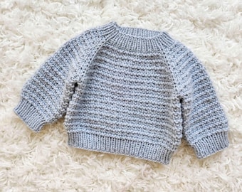 May include: A light blue knitted sweater with long sleeves. The sweater has a ribbed neckline, cuffs, and hem. The knit pattern is a textured stitch, creating a cozy appearance. The sweater is designed for a child.