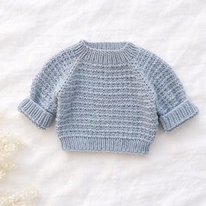 Puede incluir: Un suéter de bebé tejido en azul claro, con cuello redondo y mangas largas. Las mangas tienen puños en las muñecas. El suéter está hecho de una tela de punto texturizada y se muestra sobre una superficie blanca.