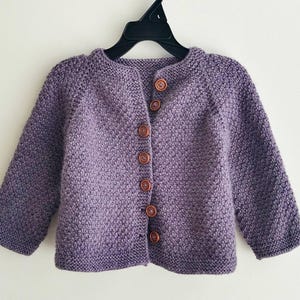 May include: A purple knitted cardigan with long sleeves and a round neckline. The button-down sweater has six brown buttons and is displayed on a black hanger. The textured knitwear is a soft, comfortable garment.
