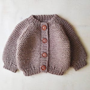 May include: A chunky knit cardigan in a muted mauve color featuring a round neckline, long sleeves with ribbed cuffs, and a front button closure with four round, dark brown wooden buttons. The garment has a textured, garter stitch pattern.